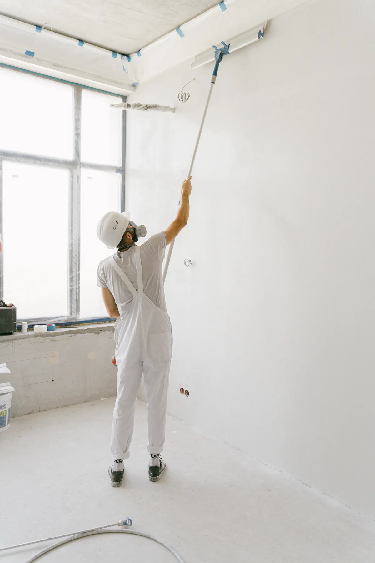 Pintor com uniforme e equipamento de proteção aplicando tinta com rolo em um ambiente interno iluminado